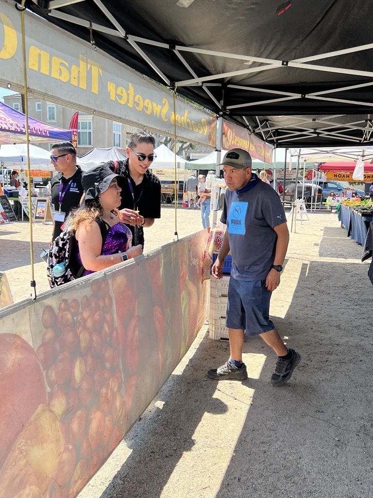 Image – 2025-08-18T112053.510 A young woman using a white cane speaks with a vendor at a farmer's market.