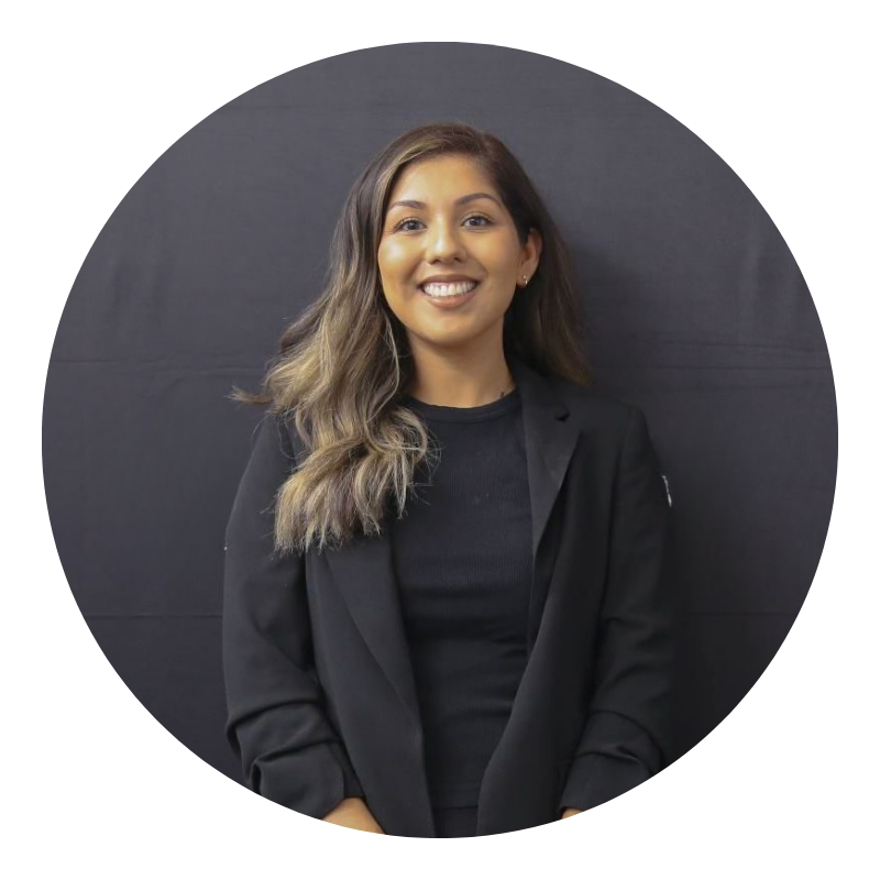A women with long dark hair, standing in front of a dark colored background, wearing a black shirt and black blazer