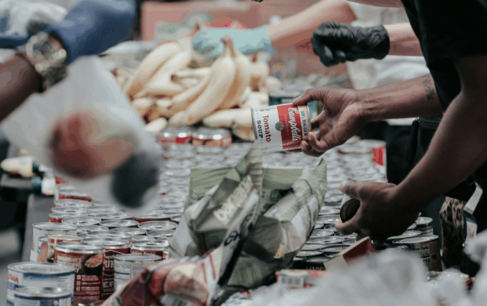 People gathered at a food pantry. Canned food items and bananas are pictures