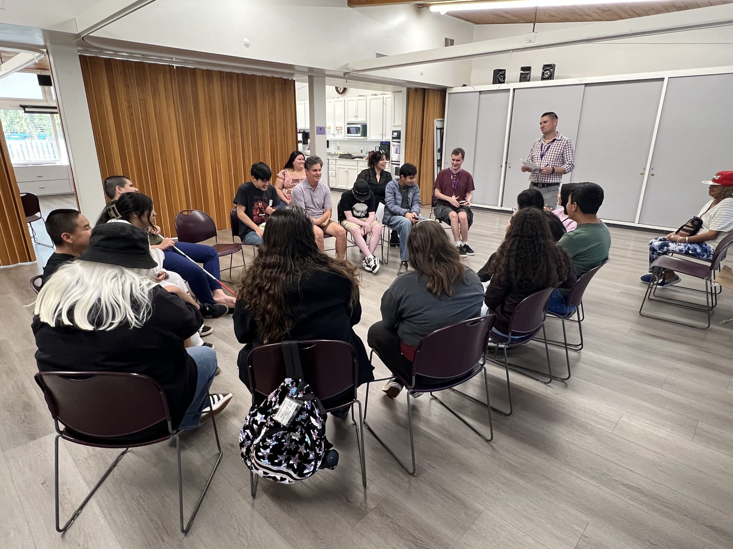 A group of people sit in a large circle inside a community room, listening to a standing speaker at the front. The room has wood-paneled dividers, light-colored walls, and a kitchen area in the background.