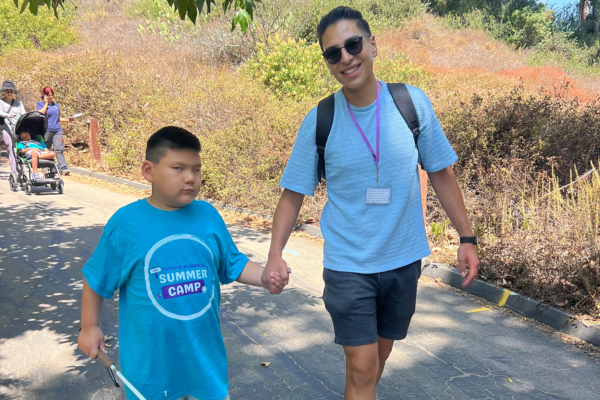 A young boy walking with a white cane, holding the hand of a volunteer