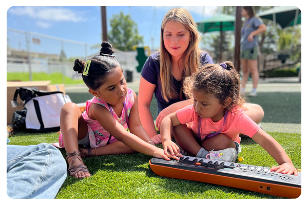 Three children and an adult sit on outdoor turf, exploring a keyboard together during a summer camp activity