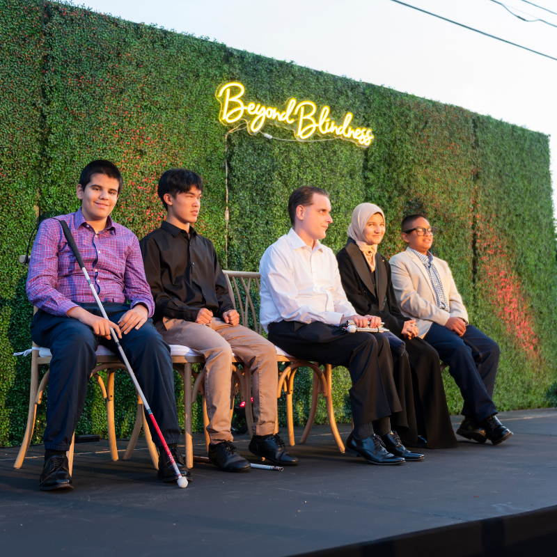 Five panelists with visual impairments sit on chairs on a stage in front of a green hedge wall with a glowing “Beyond Blindness” sign, participating in a panel discussion at a Beyond Blindness event.