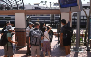 Group a visually impaired teens and young adults view the train schedule near a train track.