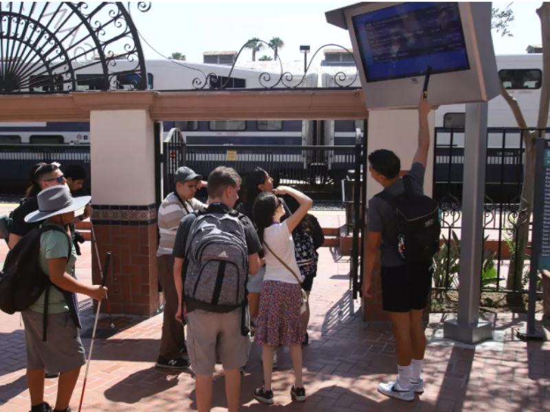 Group a visually impaired teens and young adults view the train schedule near a train track.