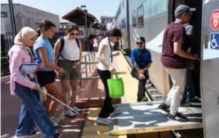 Seven people walking onto a train. One holding a white cane.
